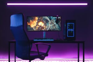 A gaming setup with a monitor, keyboard, chair, and a computer tower under purple lighting