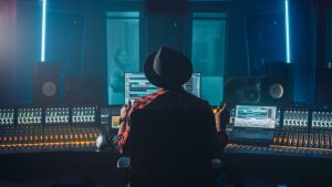 Person sitting in front of a mixing console in a music studio, wearing a hat